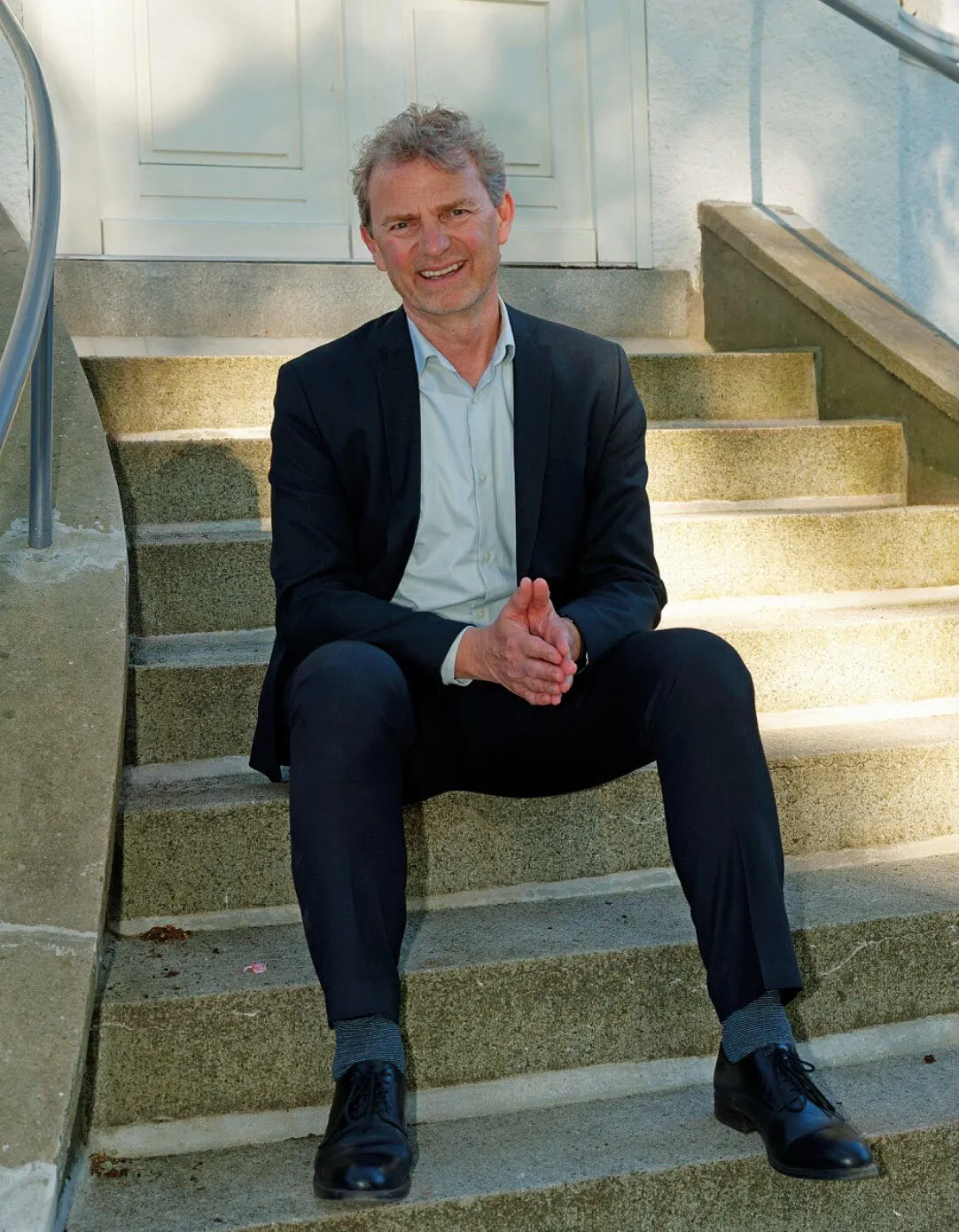 Peter Altmeppen sitzt entspannt auf einer Steintreppe vor einer hellen Tür, die Hände gefaltet.