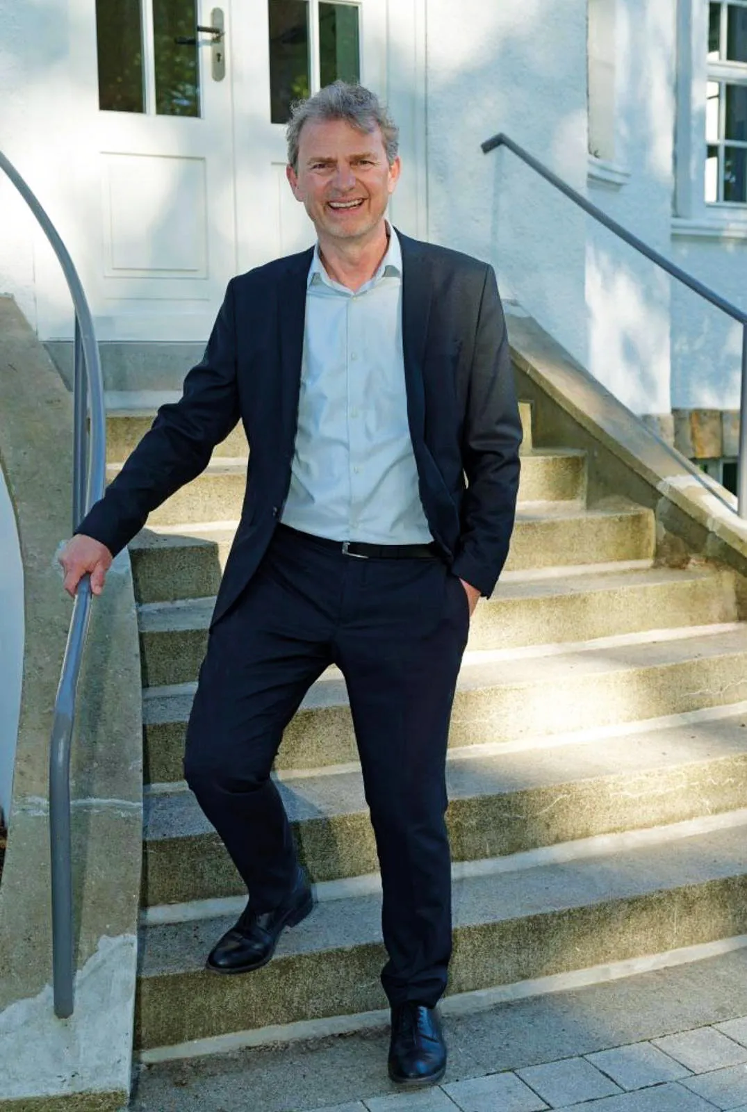 Peter Altmeppen steht lächelnd auf einer Steintreppe, im dunklen Anzug mit offenem Hemd.